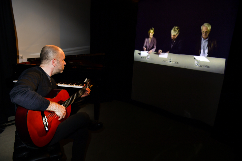 A man playing the guitar in front of a video simulator of people, reviewing his music in a dark room.